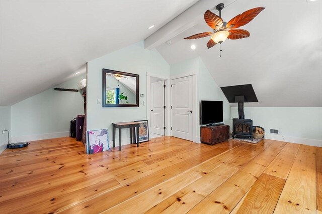 additional living space with ceiling fan, a wood stove, baseboards, wood-type flooring, and vaulted ceiling with beams