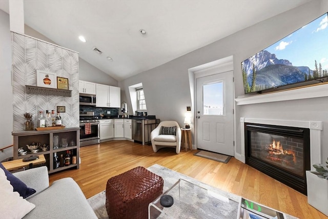 living room featuring vaulted ceiling, a glass covered fireplace, light wood-style flooring, visible vents, and recessed lighting