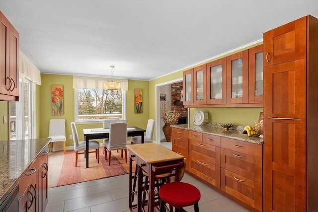 kitchen with dishwashing machine, glass insert cabinets, light stone countertops, brown cabinetry, and decorative light fixtures