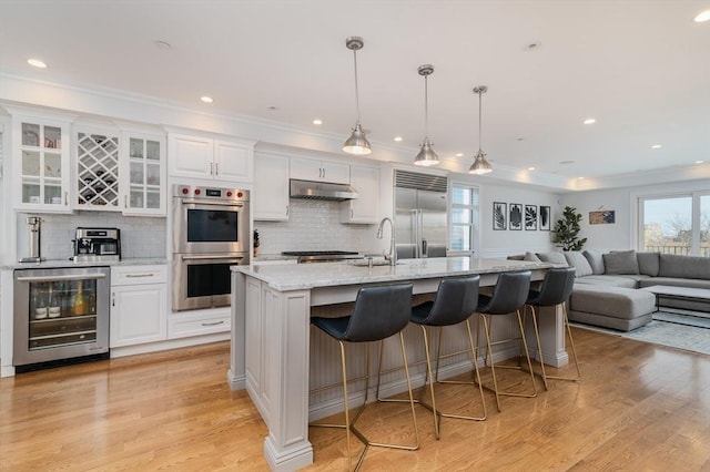 kitchen with wine cooler, glass insert cabinets, appliances with stainless steel finishes, white cabinets, and under cabinet range hood