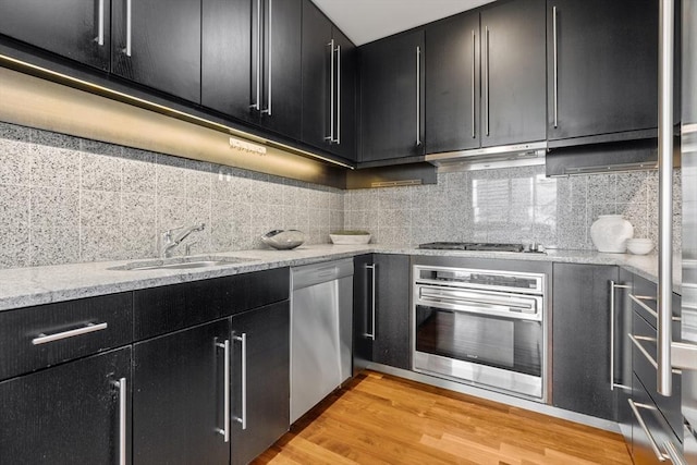 kitchen featuring under cabinet range hood, dark cabinets, stainless steel appliances, backsplash, and light wood finished floors