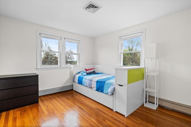 bedroom featuring a baseboard heating unit, multiple windows, wood finished floors, and visible vents