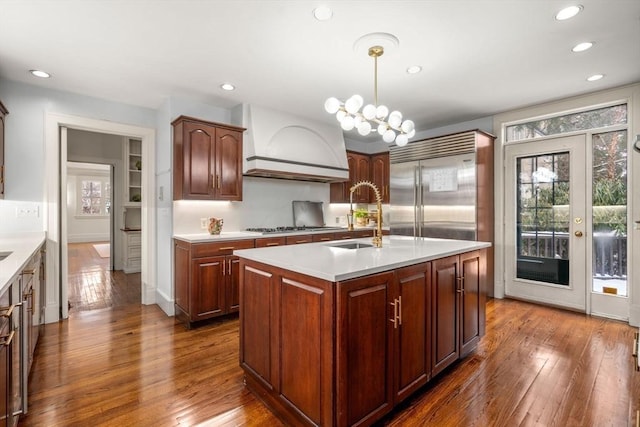 kitchen featuring a center island with sink, hanging light fixtures, premium range hood, stainless steel appliances, and sink