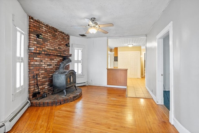 unfurnished living room featuring a baseboard heating unit, a wood stove, a ceiling fan, light wood-type flooring, and a textured ceiling