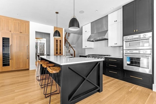 kitchen featuring decorative light fixtures, an island with sink, white cabinets, stainless steel appliances, and wall chimney exhaust hood