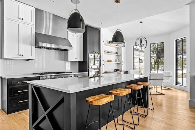 kitchen featuring wall chimney exhaust hood, hanging light fixtures, an island with sink, stainless steel appliances, and white cabinets