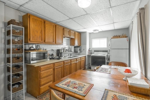 kitchen with white appliances, a drop ceiling, light countertops, decorative backsplash, and brown cabinets