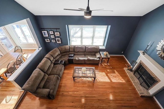 living area with a brick fireplace, a ceiling fan, baseboards, and wood finished floors