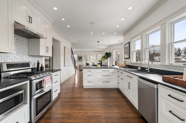 kitchen with dark countertops, appliances with stainless steel finishes, a peninsula, under cabinet range hood, and a sink