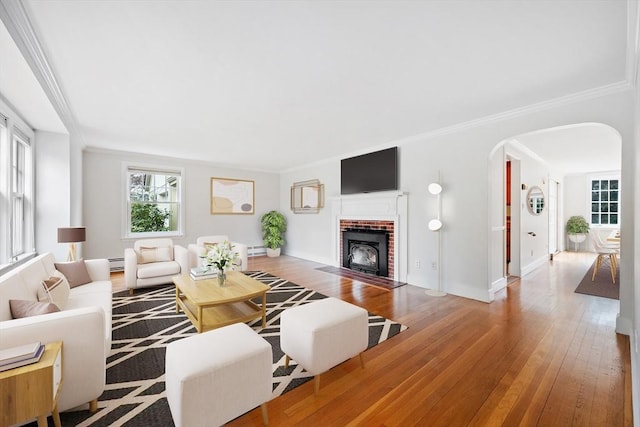 living room with ornamental molding, a brick fireplace, baseboard heating, hardwood / wood-style flooring, and arched walkways