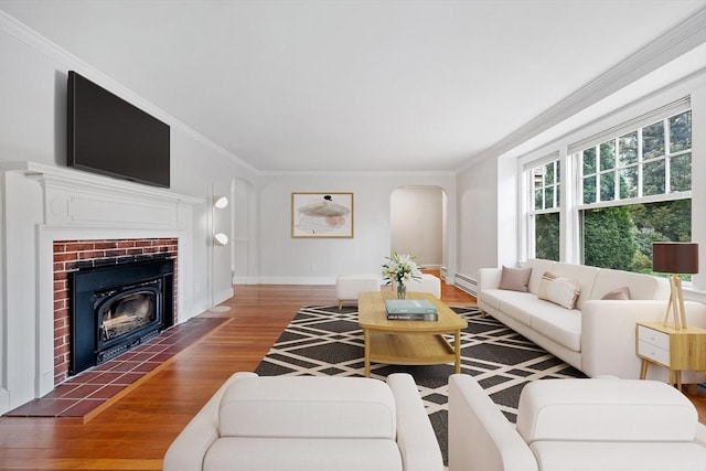 living area featuring a brick fireplace, arched walkways, wood finished floors, a baseboard radiator, and ornamental molding