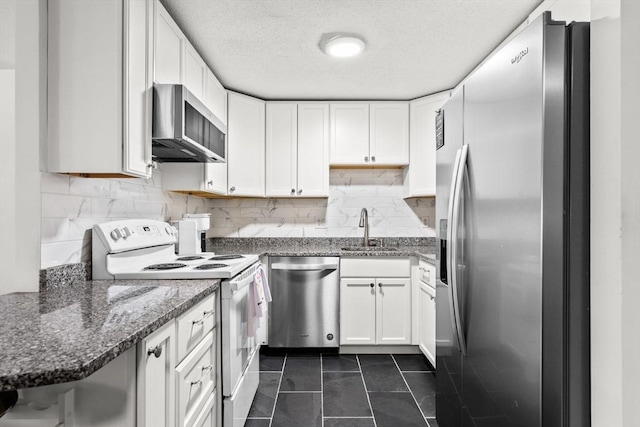 kitchen with wall chimney range hood, stainless steel appliances, white cabinets, dark tile patterned flooring, and dark stone counters