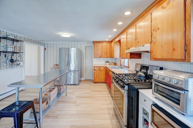 kitchen with wallpapered walls, under cabinet range hood, appliances with stainless steel finishes, light wood-style floors, and a sink