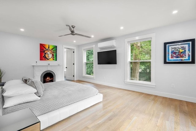 bedroom with multiple windows, light wood-style floors, a warm lit fireplace, and a wall unit AC