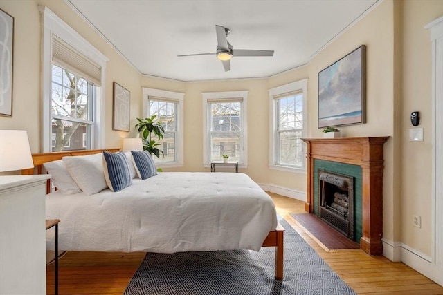 bedroom with baseboards, wood-type flooring, ceiling fan, ornamental molding, and a fireplace with flush hearth