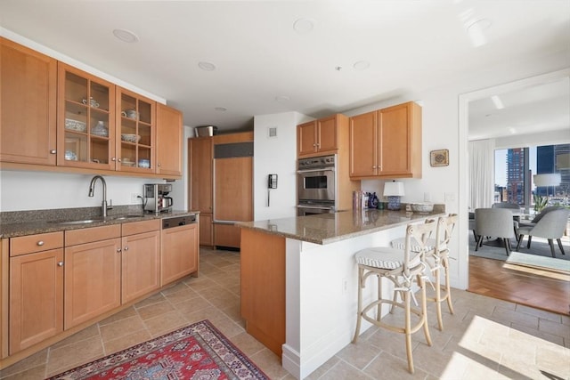 kitchen featuring double oven, dark stone countertops, a sink, glass insert cabinets, and a breakfast bar area