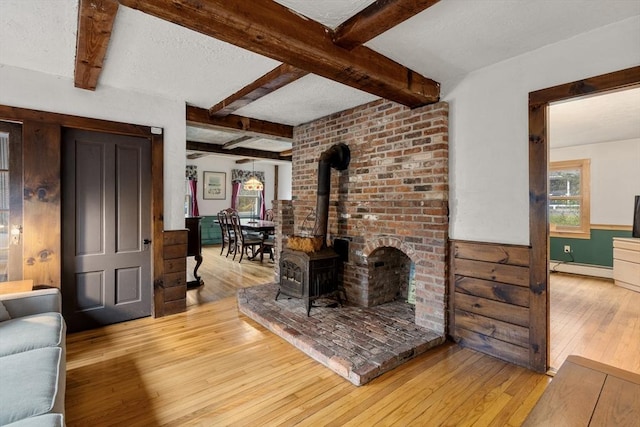 living area with a wood stove, a wainscoted wall, beamed ceiling, a baseboard heating unit, and hardwood / wood-style flooring