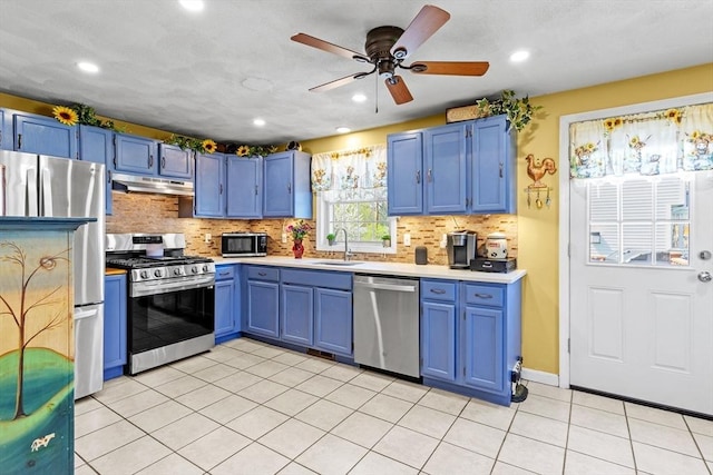 kitchen with blue cabinetry, appliances with stainless steel finishes, a sink, light countertops, and under cabinet range hood