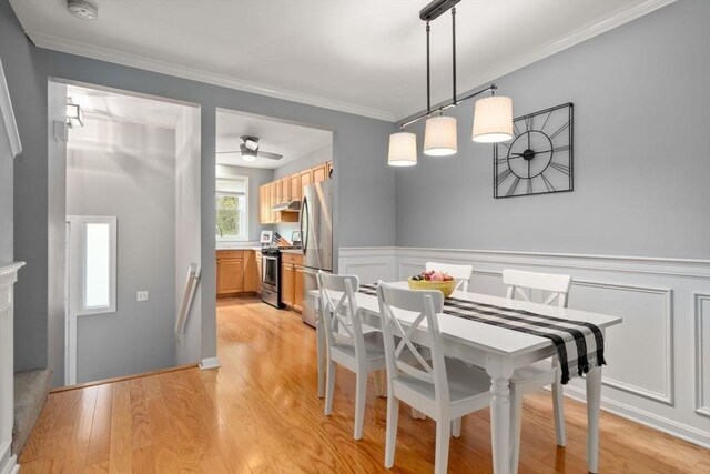 dining space with crown molding, ceiling fan, a decorative wall, a wainscoted wall, and light wood-type flooring