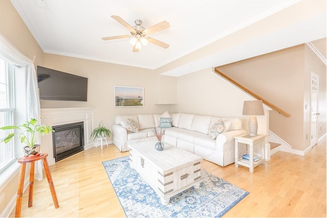 living area with crown molding, ceiling fan, a glass covered fireplace, light wood-style flooring, and baseboards