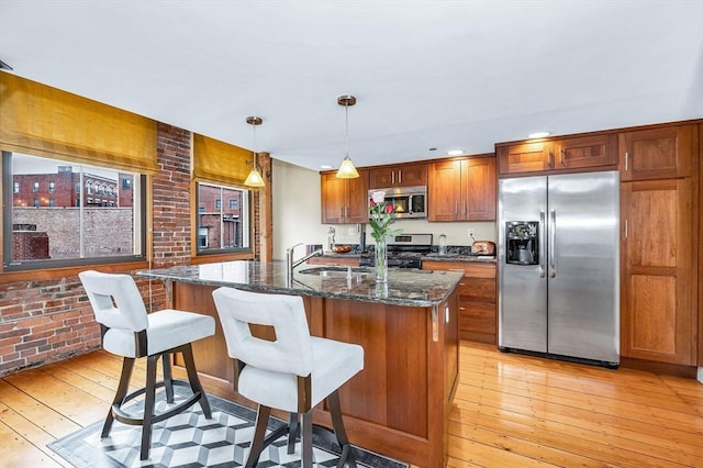 kitchen with brown cabinetry, appliances with stainless steel finishes, light wood-type flooring, dark stone countertops, and an island with sink