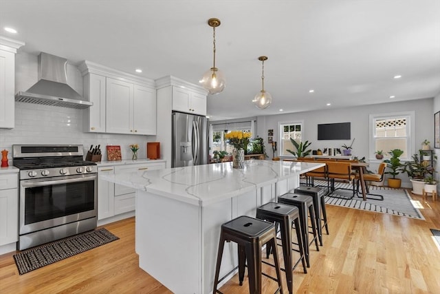 kitchen with appliances with stainless steel finishes, white cabinetry, wall chimney range hood, a kitchen island, and light hardwood / wood-style flooring