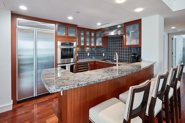 kitchen with tasteful backsplash, appliances with stainless steel finishes, kitchen peninsula, light stone countertops, and exhaust hood