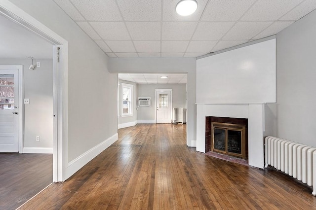 unfurnished living room with radiator, a drop ceiling, dark wood-type flooring, and a wall mounted air conditioner