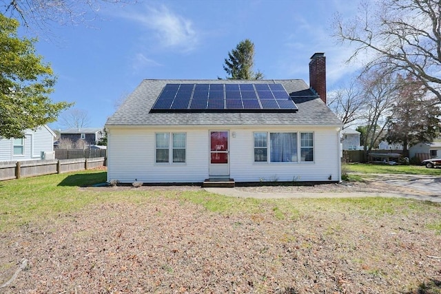 back of property featuring roof mounted solar panels, a shingled roof, a chimney, and fence