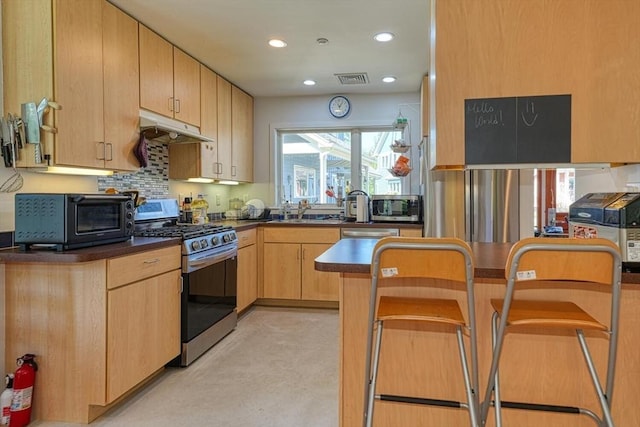 kitchen with stainless steel appliances, under cabinet range hood, dark countertops, light brown cabinets, and concrete floors