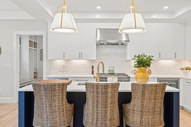 kitchen featuring decorative light fixtures, a center island with sink, light countertops, a tray ceiling, and wall chimney exhaust hood