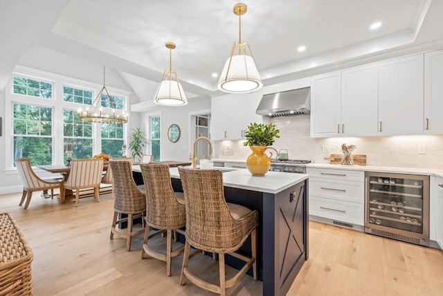 kitchen featuring a tray ceiling, decorative backsplash, light countertops, wine cooler, and wall chimney range hood