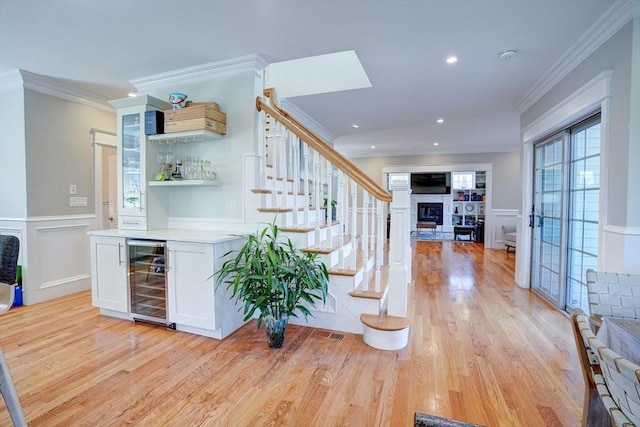 indoor dry bar featuring wine cooler, light wood-type flooring, a decorative wall, a glass covered fireplace, and crown molding