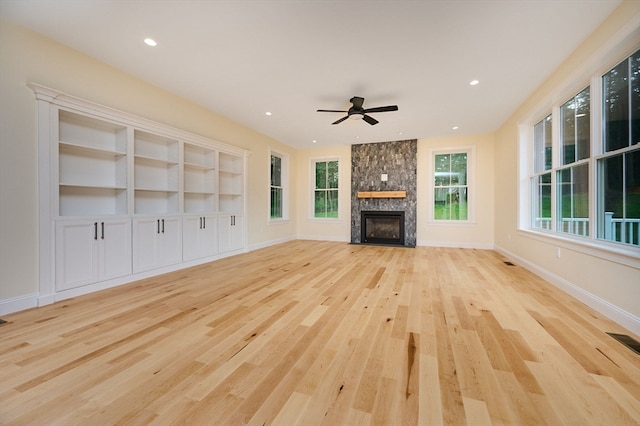 unfurnished living room featuring built in features, light hardwood / wood-style flooring, ceiling fan, and a stone fireplace