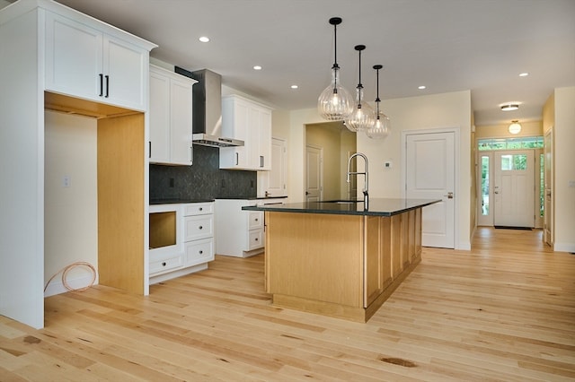 kitchen with wall chimney range hood, light wood-type flooring, a kitchen island with sink, and white cabinetry