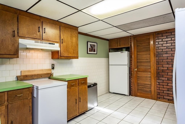 kitchen featuring brown cabinets, stove, freestanding refrigerator, light tile patterned flooring, and under cabinet range hood