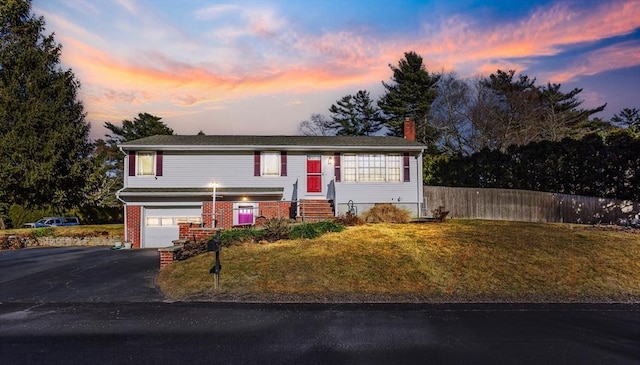 raised ranch with aphalt driveway, an attached garage, fence, a yard, and brick siding