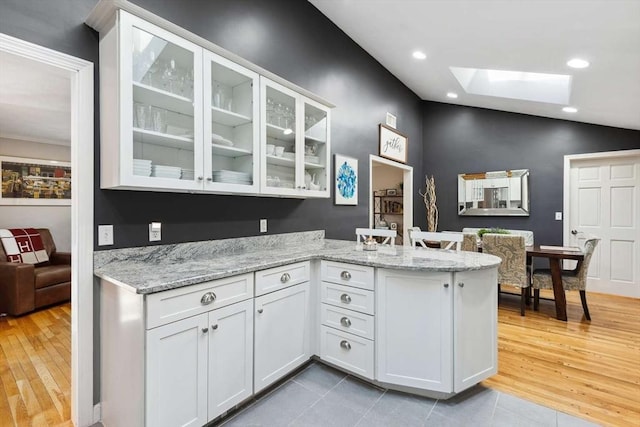 kitchen featuring light tile patterned floors, kitchen peninsula, vaulted ceiling with skylight, white cabinets, and light stone counters