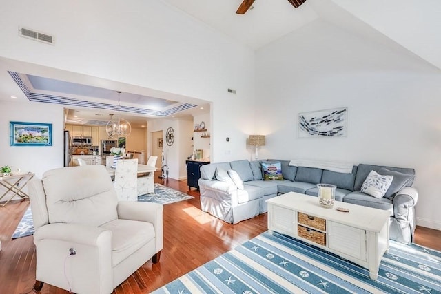 living room with a tray ceiling, high vaulted ceiling, ceiling fan with notable chandelier, and light wood-type flooring