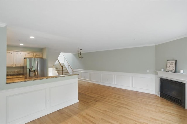unfurnished living room featuring crown molding, stairs, a glass covered fireplace, a wainscoted wall, and light wood finished floors