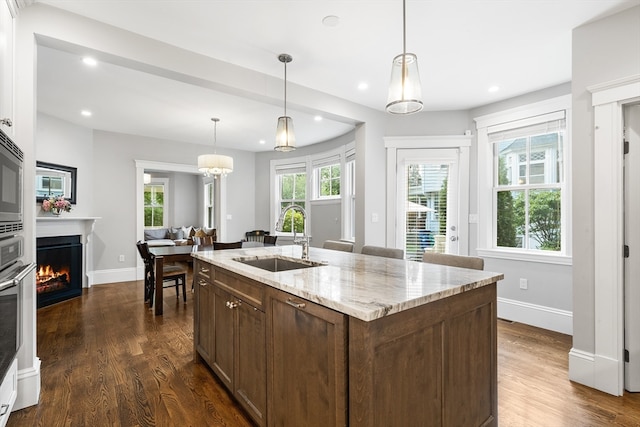 kitchen featuring hanging light fixtures, dark wood-type flooring, an island with sink, light stone countertops, and sink