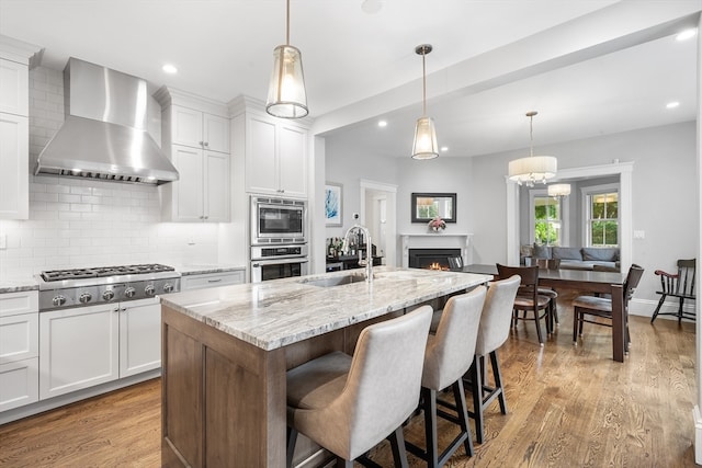 kitchen featuring white cabinets, a center island with sink, sink, and wall chimney range hood