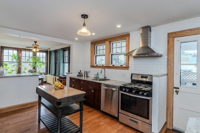 kitchen with light stone countertops, stainless steel appliances, light wood finished floors, a sink, and wall chimney exhaust hood
