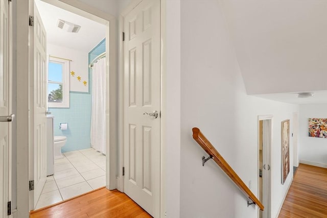hallway featuring tile walls, light wood finished floors, visible vents, a wainscoted wall, and an upstairs landing