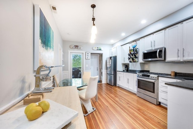 kitchen with white cabinets, appliances with stainless steel finishes, dark stone counters, hanging light fixtures, and light hardwood / wood-style flooring