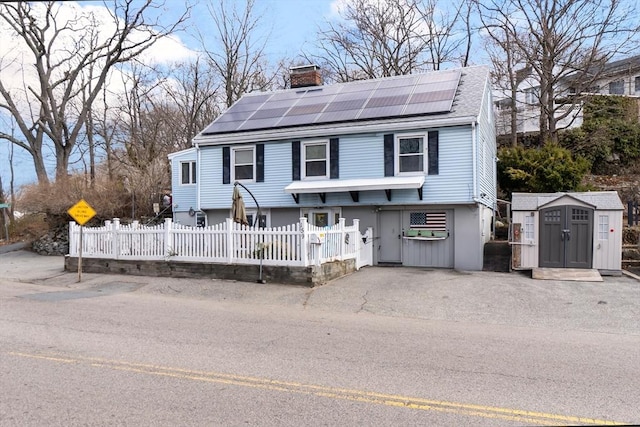 view of front facade featuring a fenced front yard, a chimney, an outdoor structure, roof mounted solar panels, and a shingled roof
