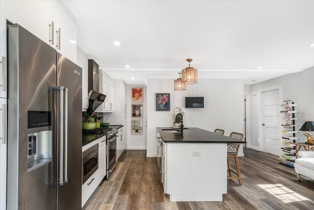 kitchen with dark countertops, high quality appliances, white cabinetry, wall chimney exhaust hood, and a sink