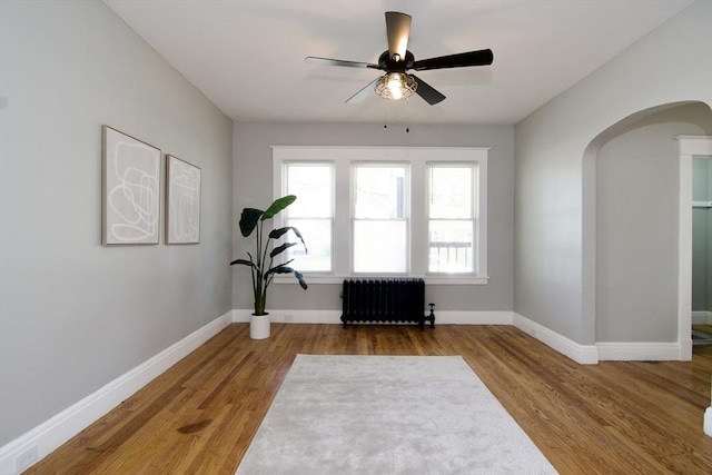 living area with arched walkways, wood finished floors, radiator heating unit, baseboards, and a ceiling fan