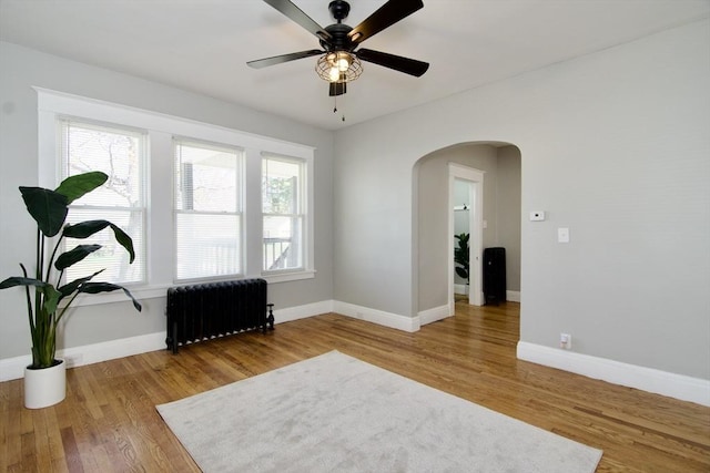 spare room featuring arched walkways, baseboards, radiator, wood finished floors, and a ceiling fan