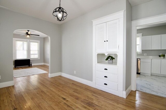 unfurnished dining area with light wood-type flooring, a ceiling fan, radiator heating unit, baseboards, and arched walkways